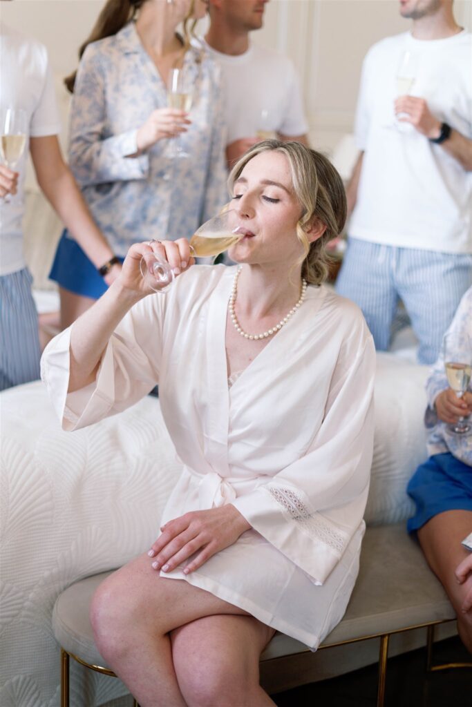 Romy sipping Champagne in a bridal robe while her friends laugh behind her