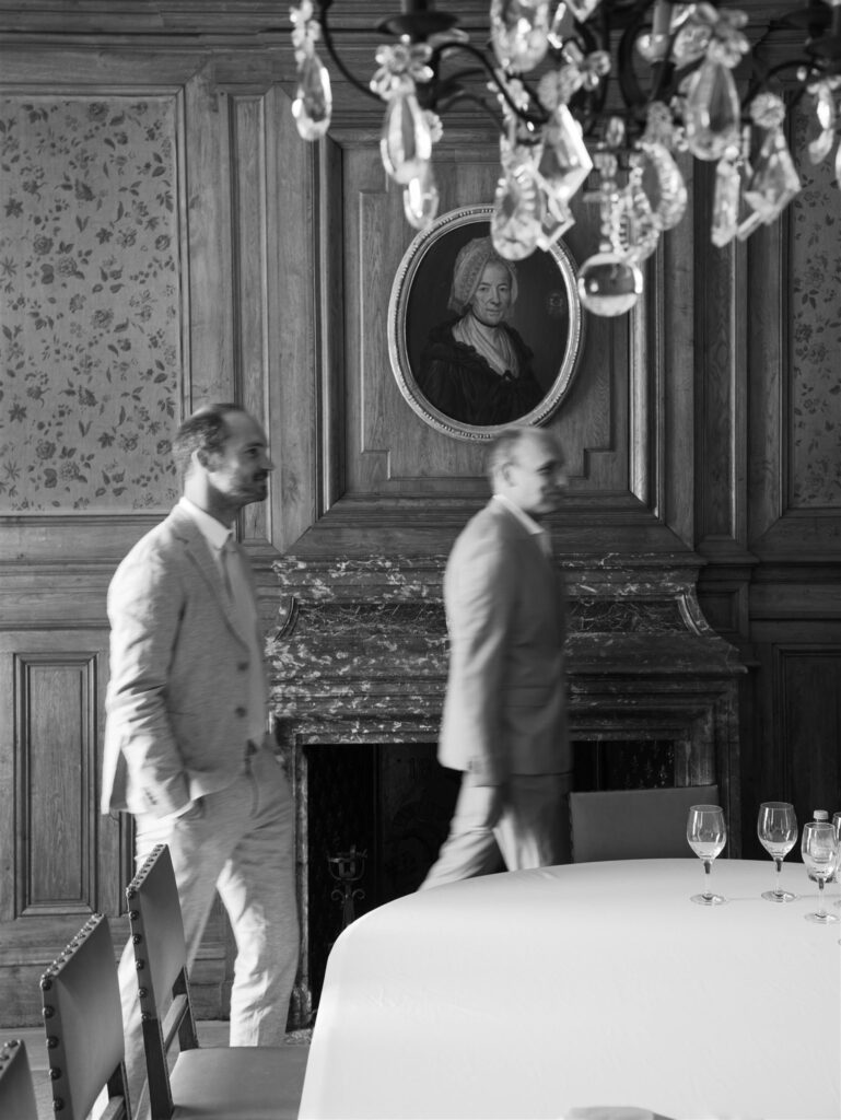 Black and white photo of two men beside a fireplace under a chandelier at the château
