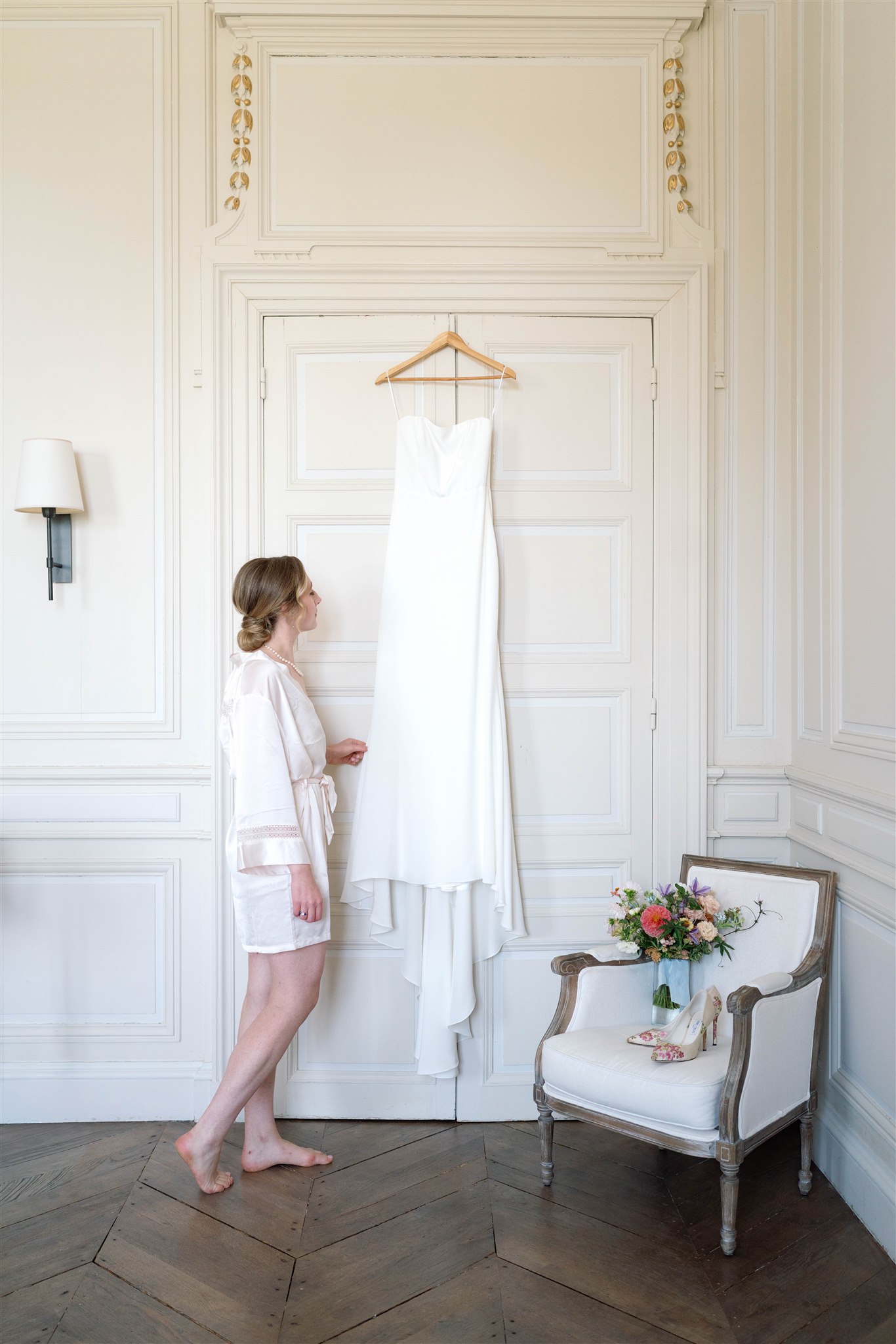 Romy in a bridal robe standing beside her wedding dress hanging on a door at the château