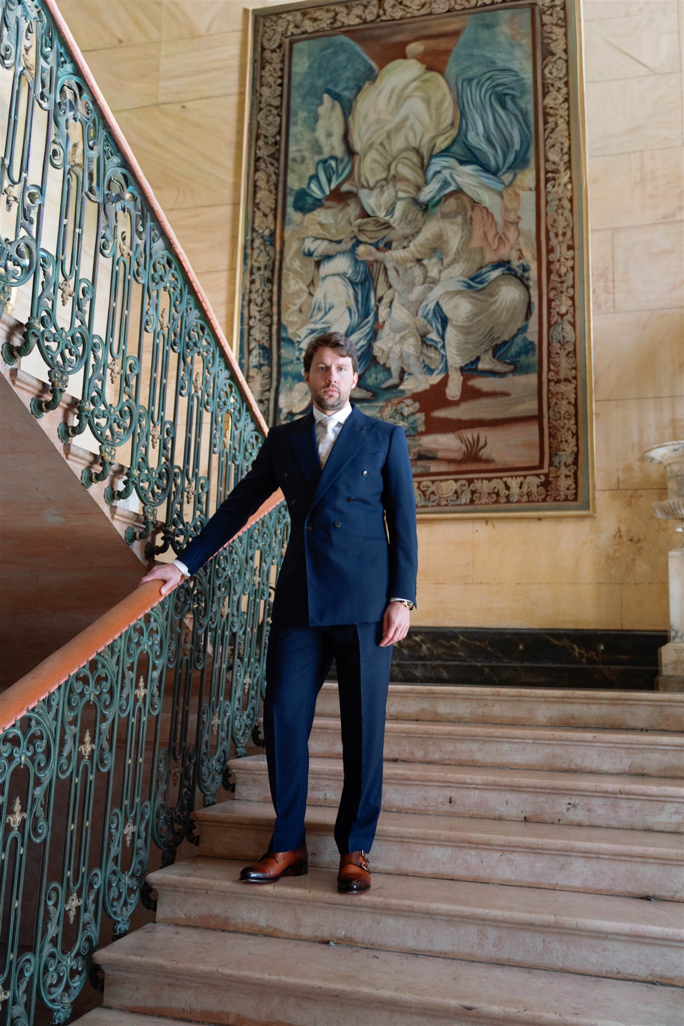 Jonathan in a navy suit standing on the grand staircase of Château de Varennes beneath a tapestry