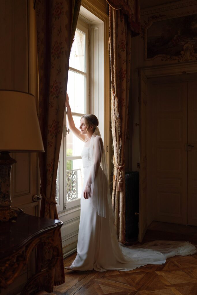 Romy gazing out a window in her wedding gown and veil in warm morning light