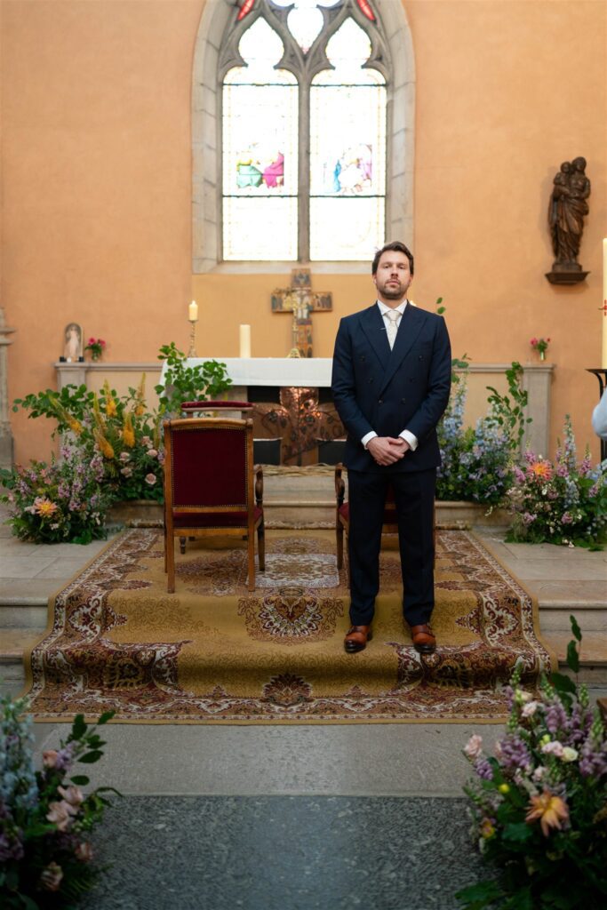 Jonathan waiting at the altar of the small church, framed by flower arrangements and stained glass