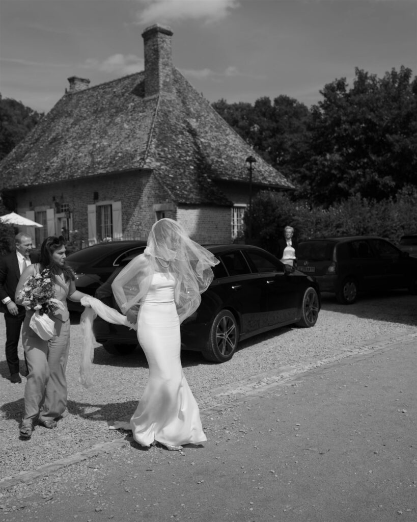 Black and white photo of Romy in her gown and veil beside a car near a thatched-roof cottage