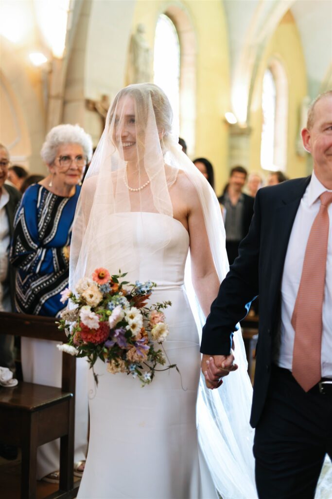 Romy walking down the aisle holding her bouquet as guests look on