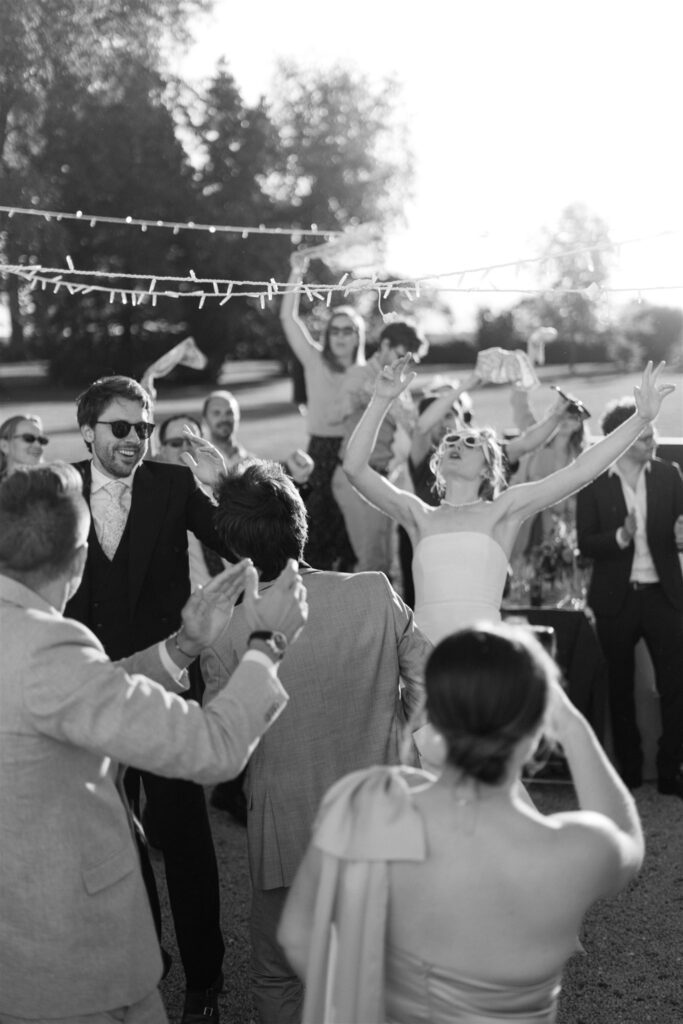 Black and white photo of guests dancing with their arms raised beneath string lights