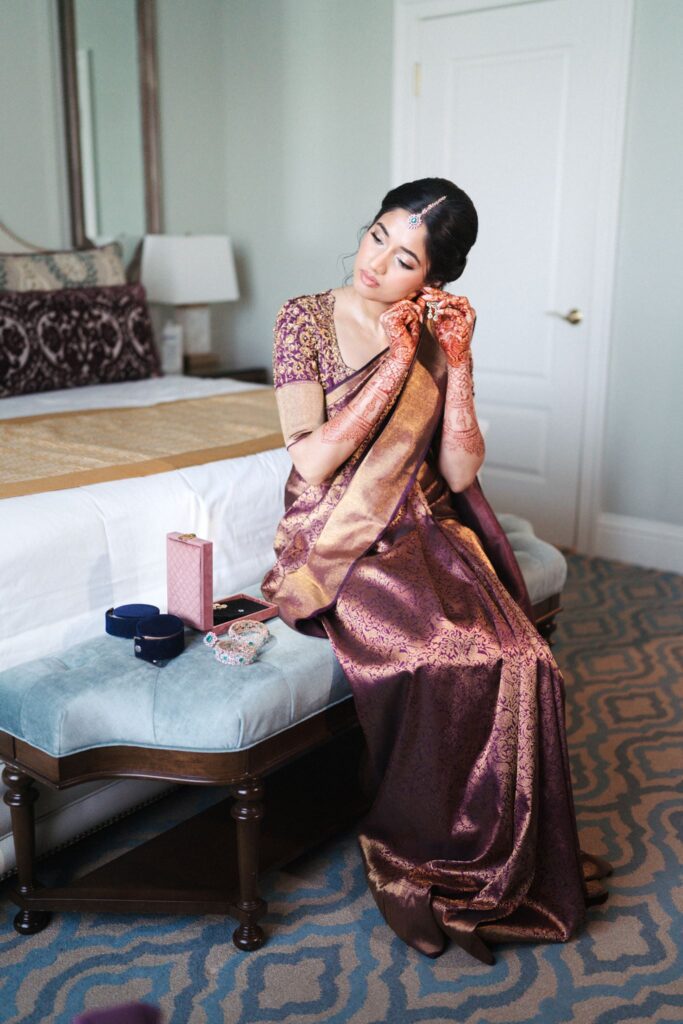 Bride in a purple and gold saree finishing her jewelry on a bench in her suite at The Biltmore Hotel