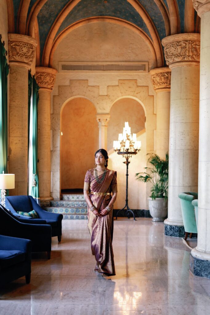 Bride in a saree standing in an arched hotel lobby with a chandelier
