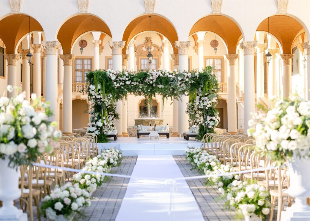 Mandap of greenery and white flowers framed by the arched loggia and aisle