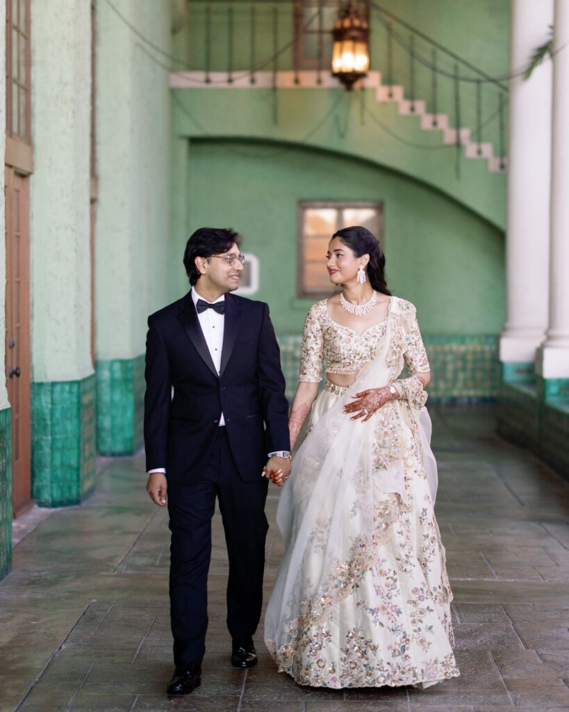 Advaitaa and Tejus holding hands in the green-tiled corridor, she in a lehenga choli and he in a tuxedo