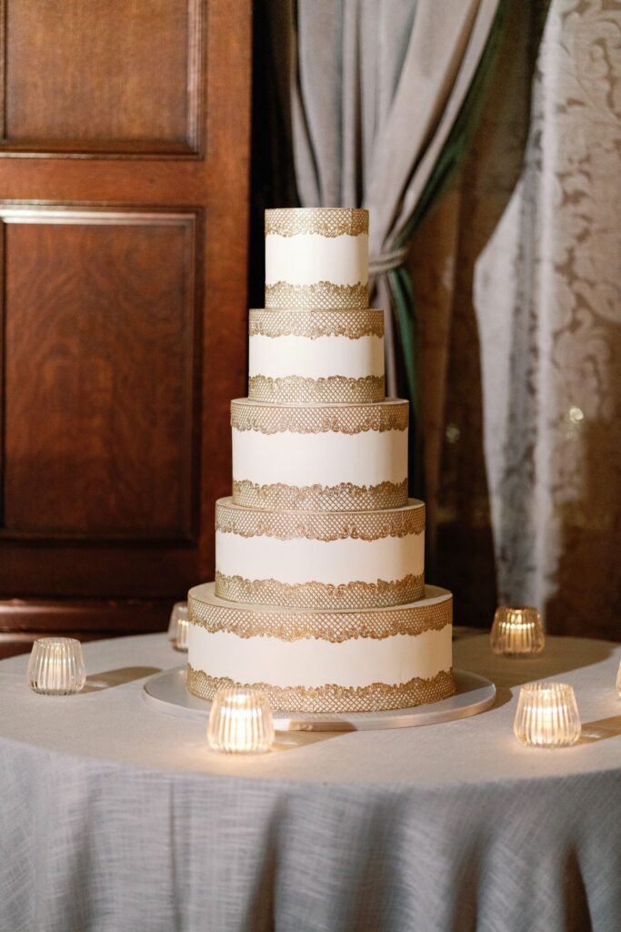Five-tier white wedding cake with gold lace detailing on a candlelit table