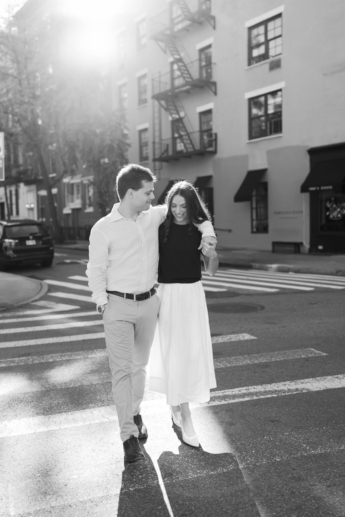 Black and White photo of Haley and Jeremy walking toward the camera with golden sun flaring behind them on a West Village street in New York City