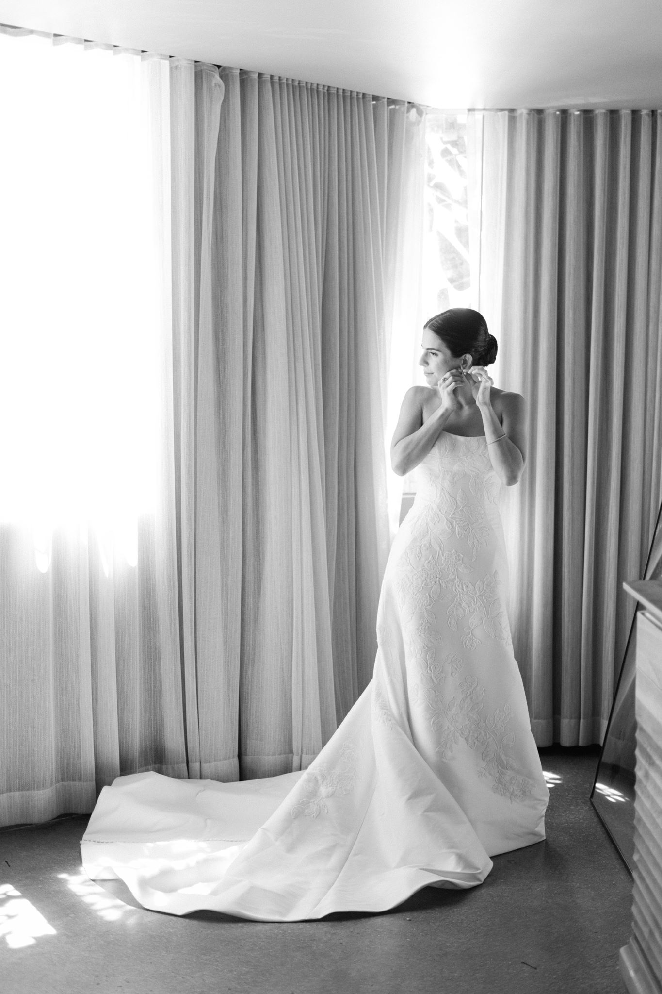 Ashley in her wedding gown adjusting her earring by a sunlit window