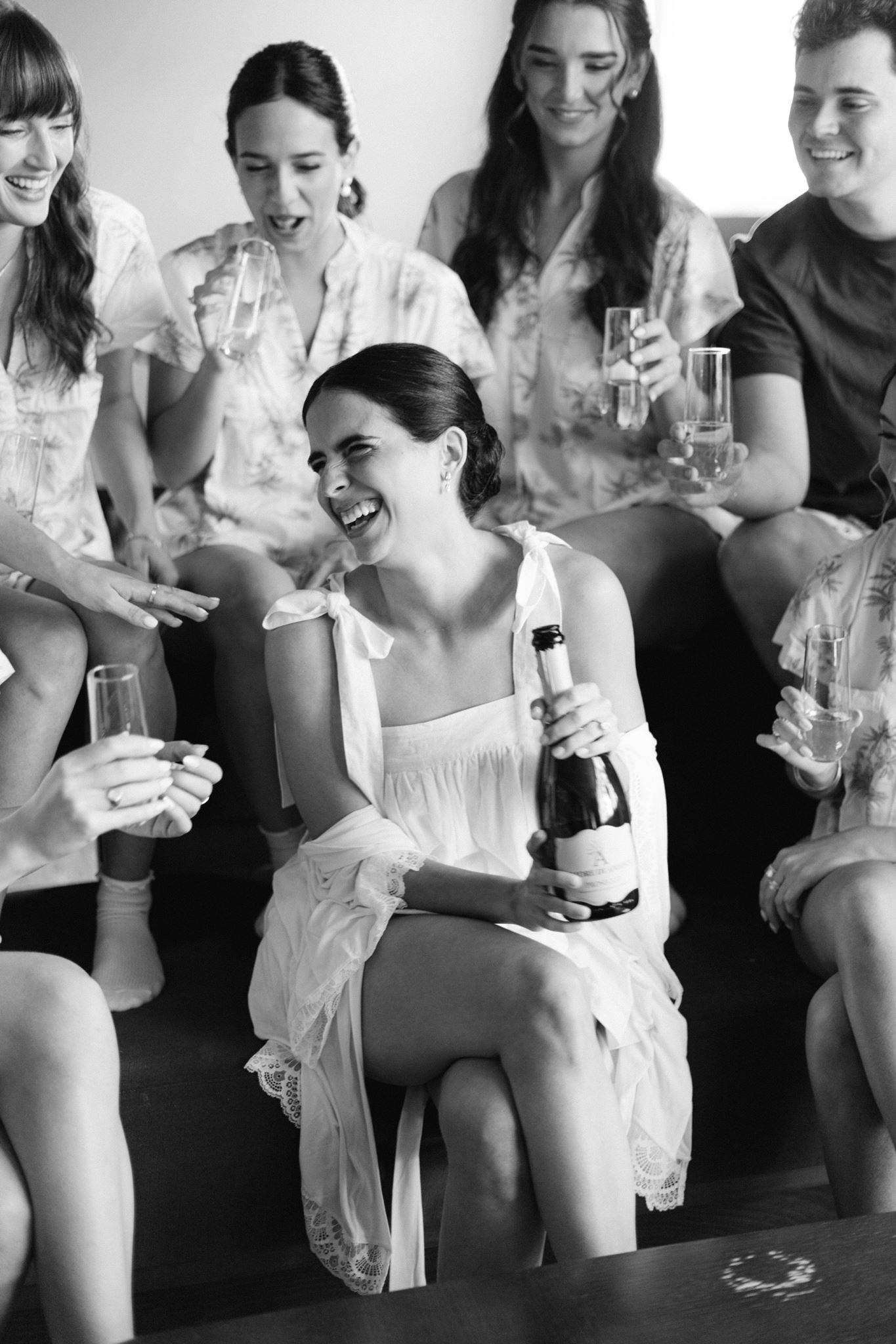 Ashley laughing in a white robe with a bottle of champagne surrounded by her bridesmaids