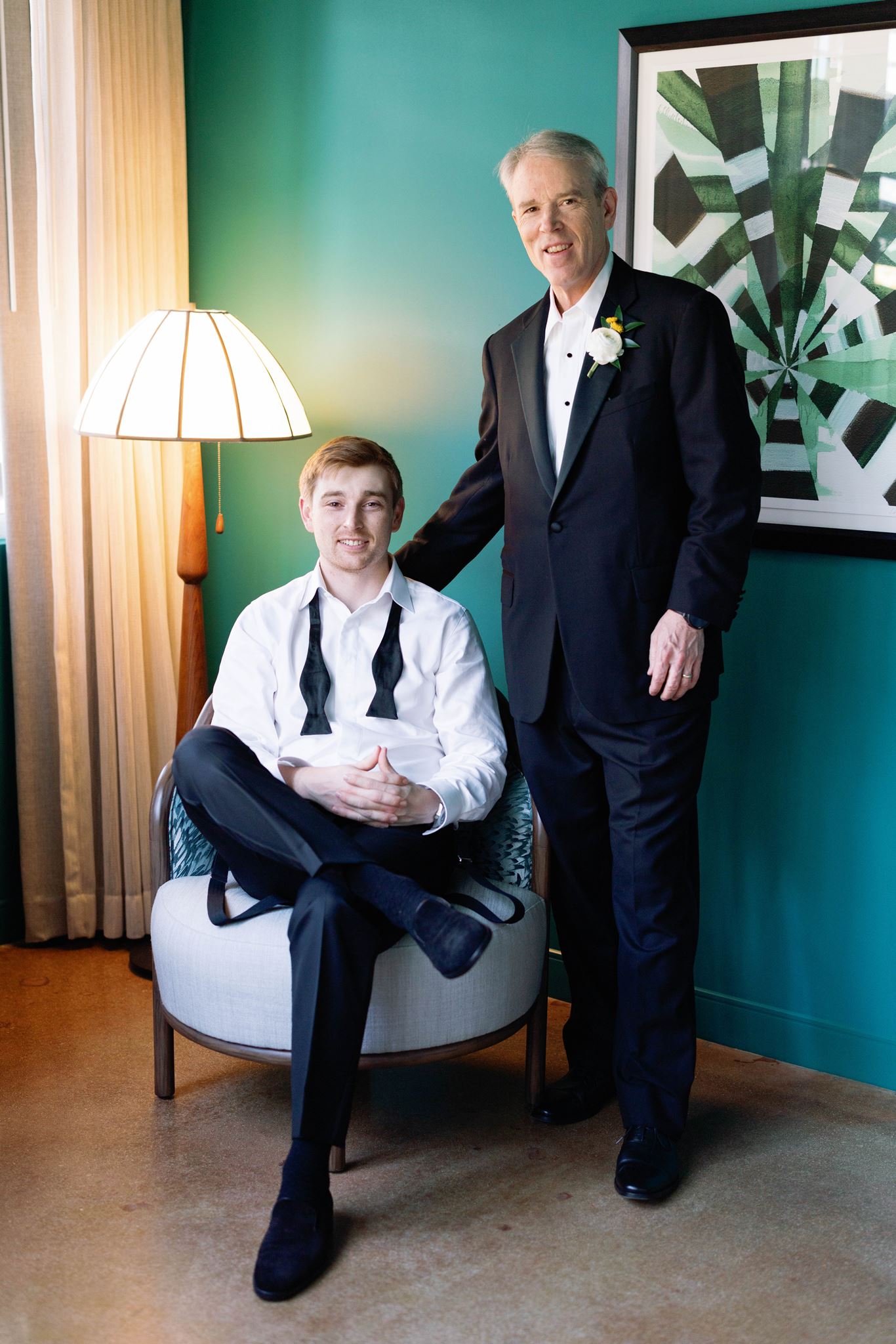 Luke seated in a teal hotel room with his father standing beside him