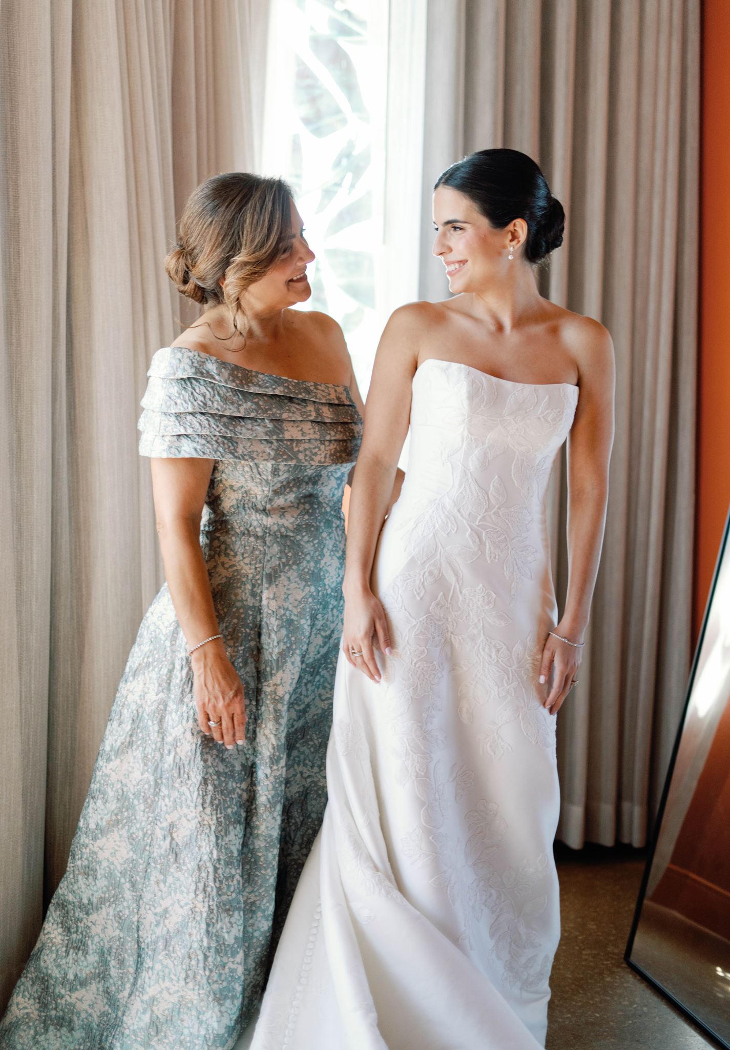 Ashley's mother helping her button her wedding gown by the window