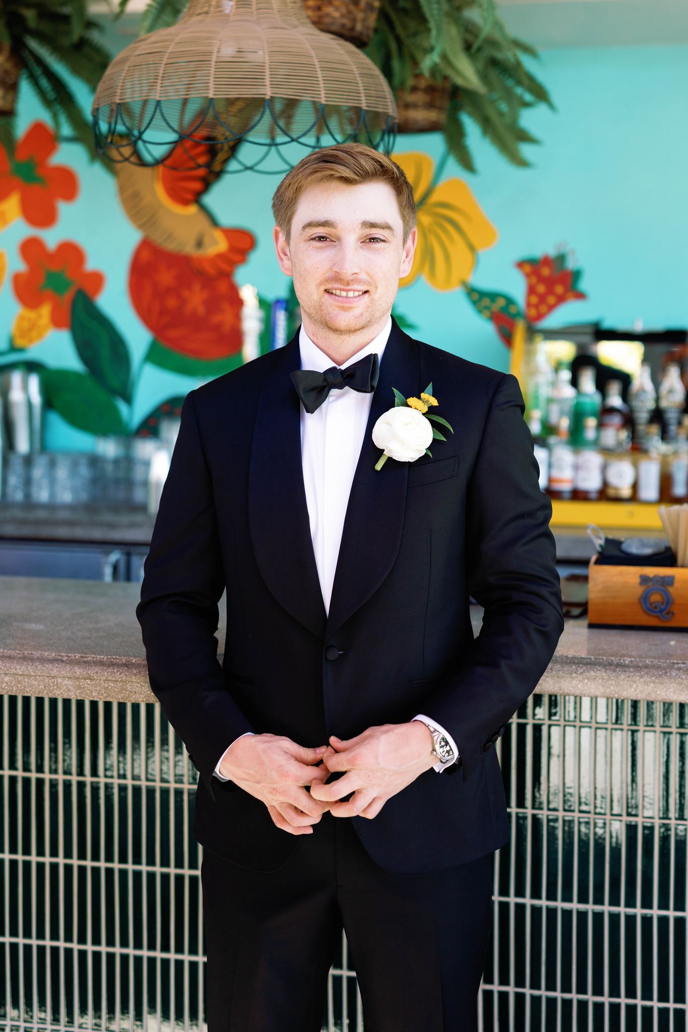 Portrait of Luke smiling in his tuxedo with a white boutonniere in front of a colorful tropical mural
