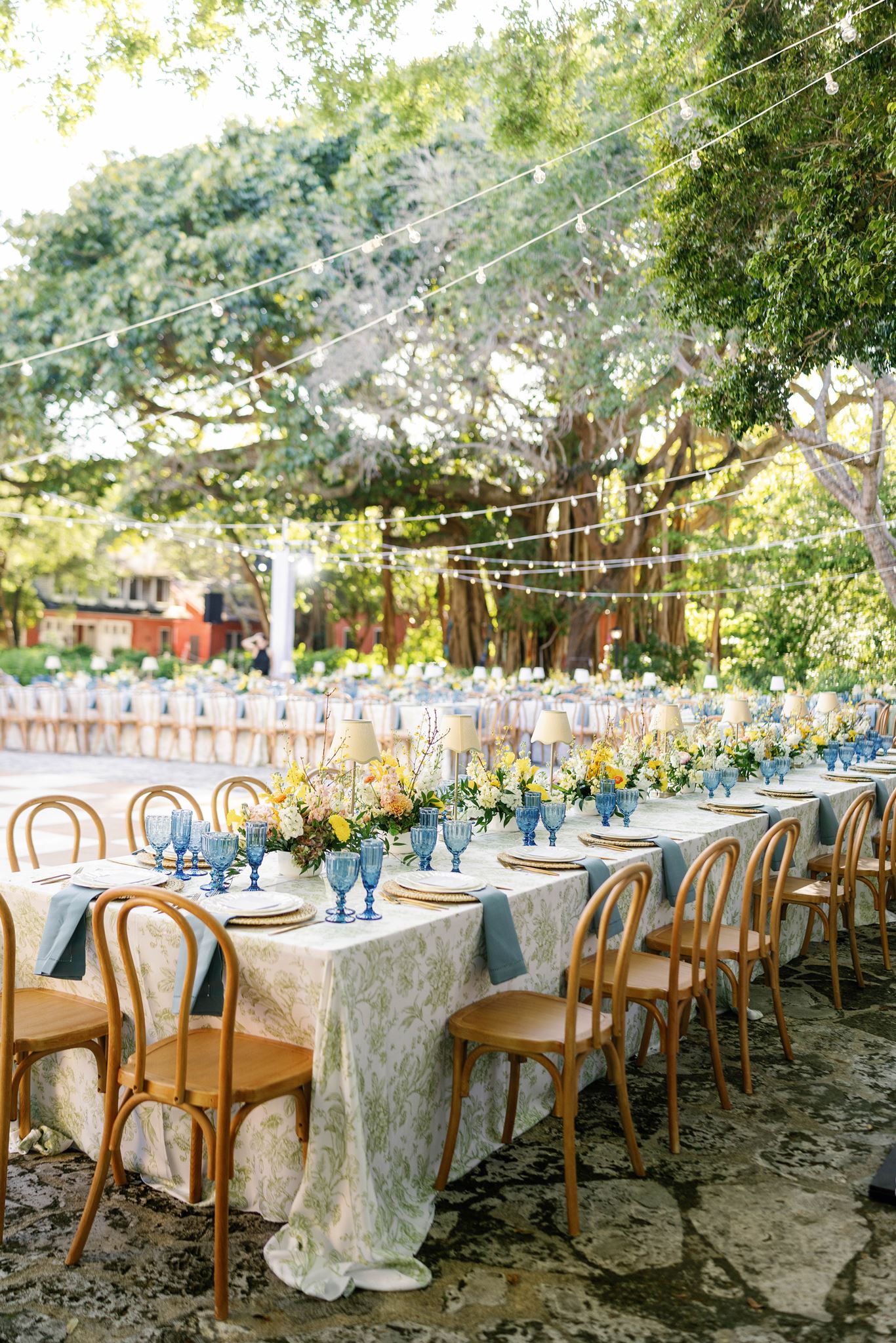 Long reception tables with green floral linens and wooden chairs under a banyan tree
