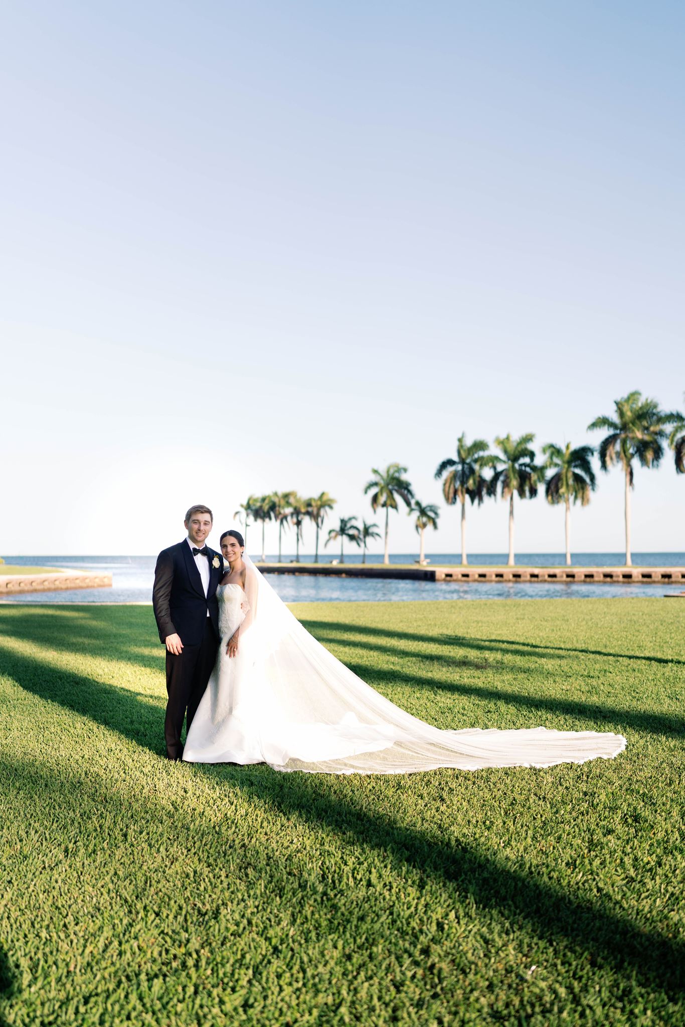 Ashley and Luke standing on the lawn beside Biscayne Bay with her cathedral veil trailing