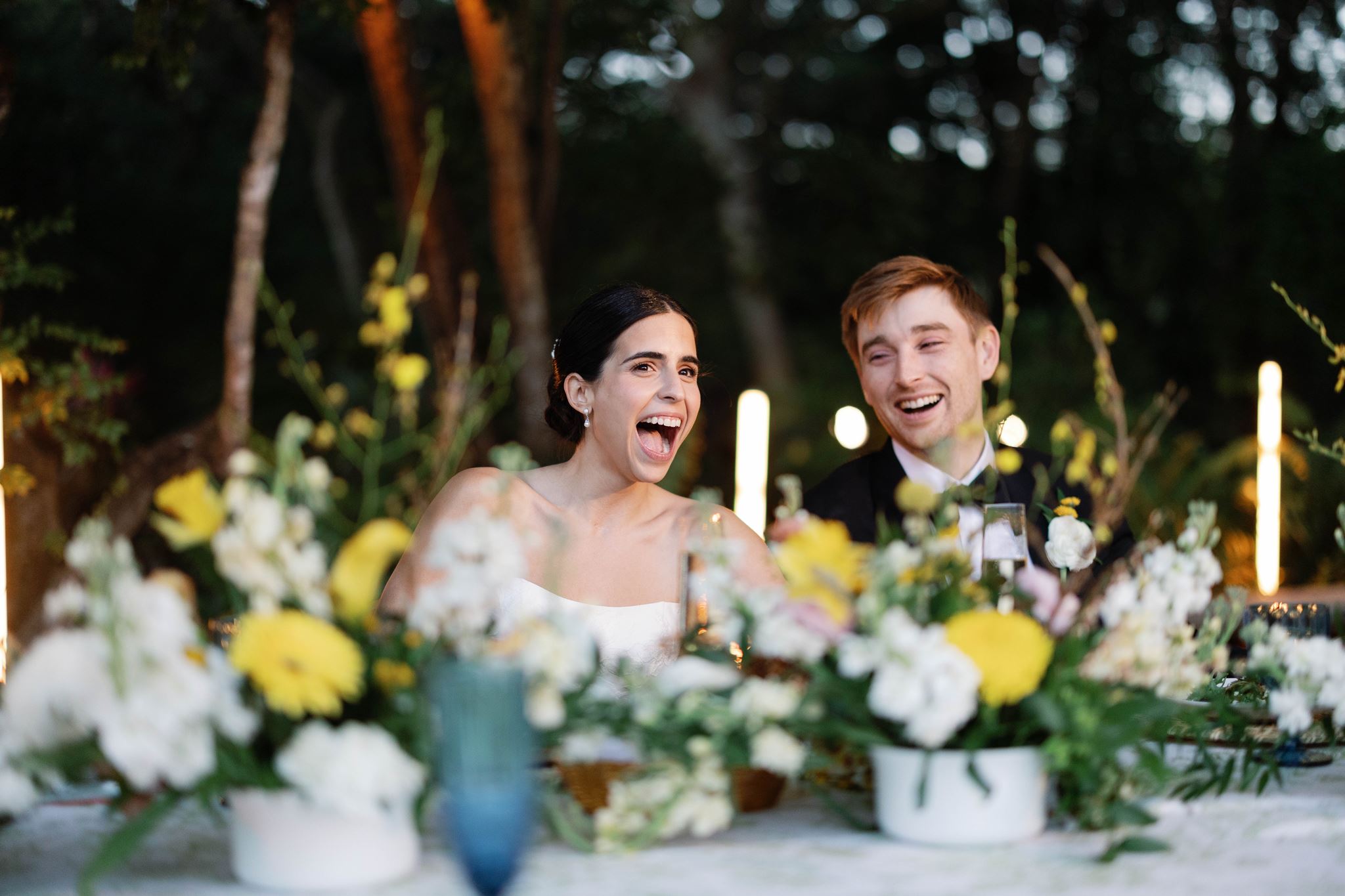 Ashley and Luke laughing at their sweetheart table surrounded by flowers