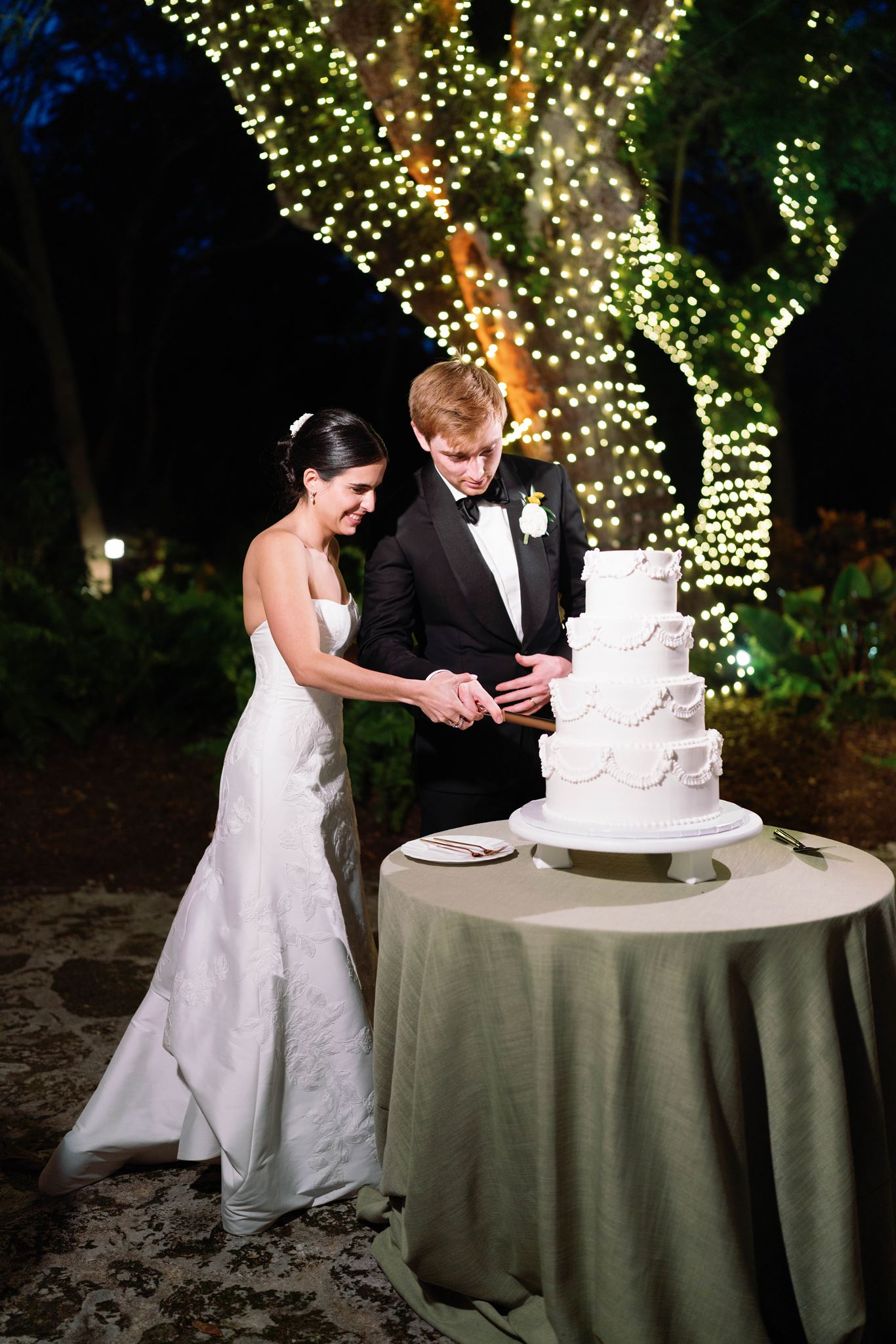 Ashley and Luke cutting their wedding cake under trees lit with string lights