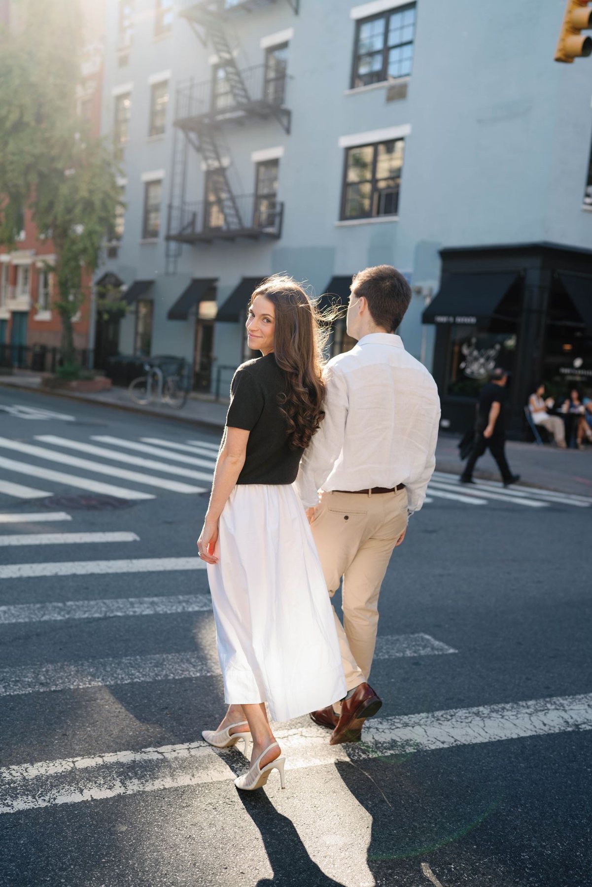 Haley and Jeremy walking hand in hand across a sunlit West Village crosswalk in New York City, Haley glancing back over her shoulder