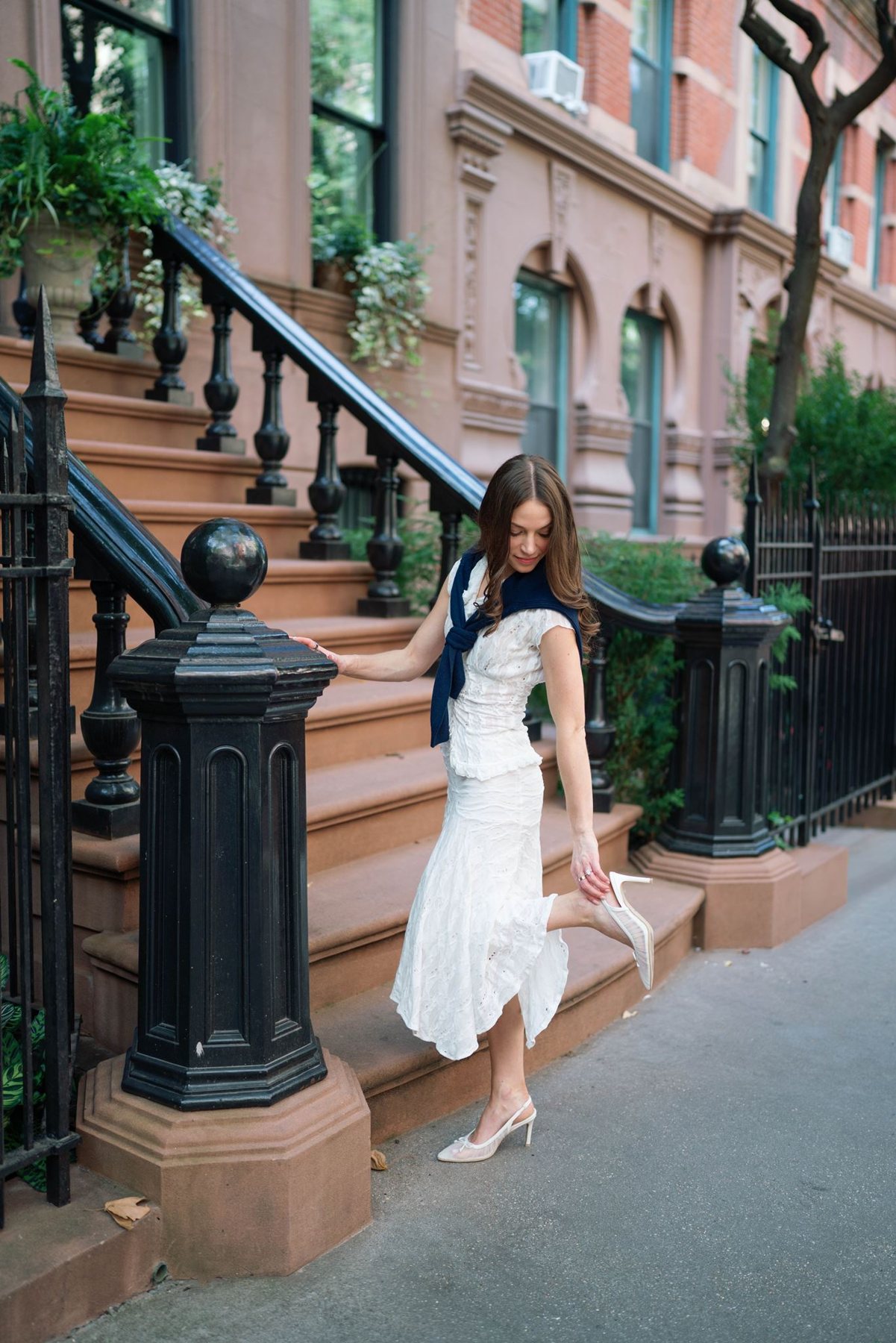 Haley in a white dress standing by the black iron stair railing of a West Village brownstone